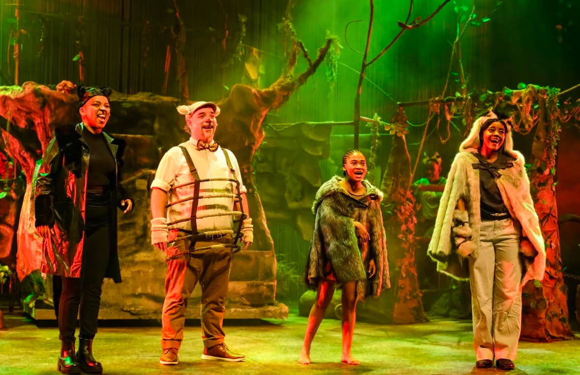 Actors portraying Bagheera, Baloo, Mowgli, and Raksha in The Jungle Book stage production, dressed in modern, stylised costumes with a jungle-themed set in the background.