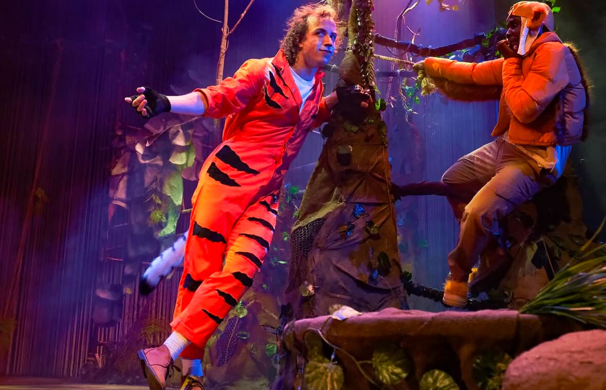 Jonathan Raath as Shere Khan in The Jungle Book stage production, wearing an orange tiger-striped jumpsuit, interacting with another actor in an immersive jungle-themed set.